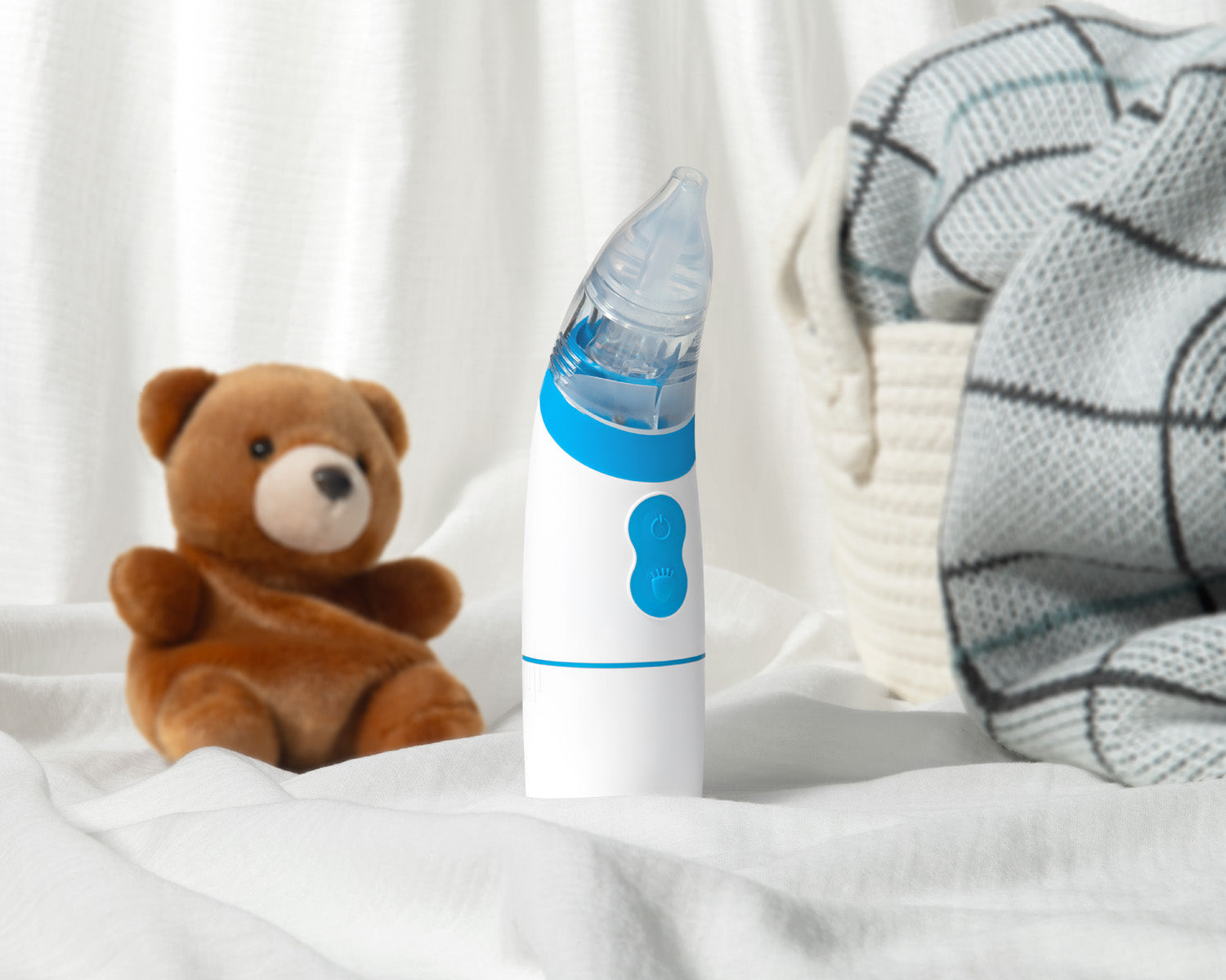 Baby nasal aspirator next to a teddy bear on a white surface with a soft blanket in the background