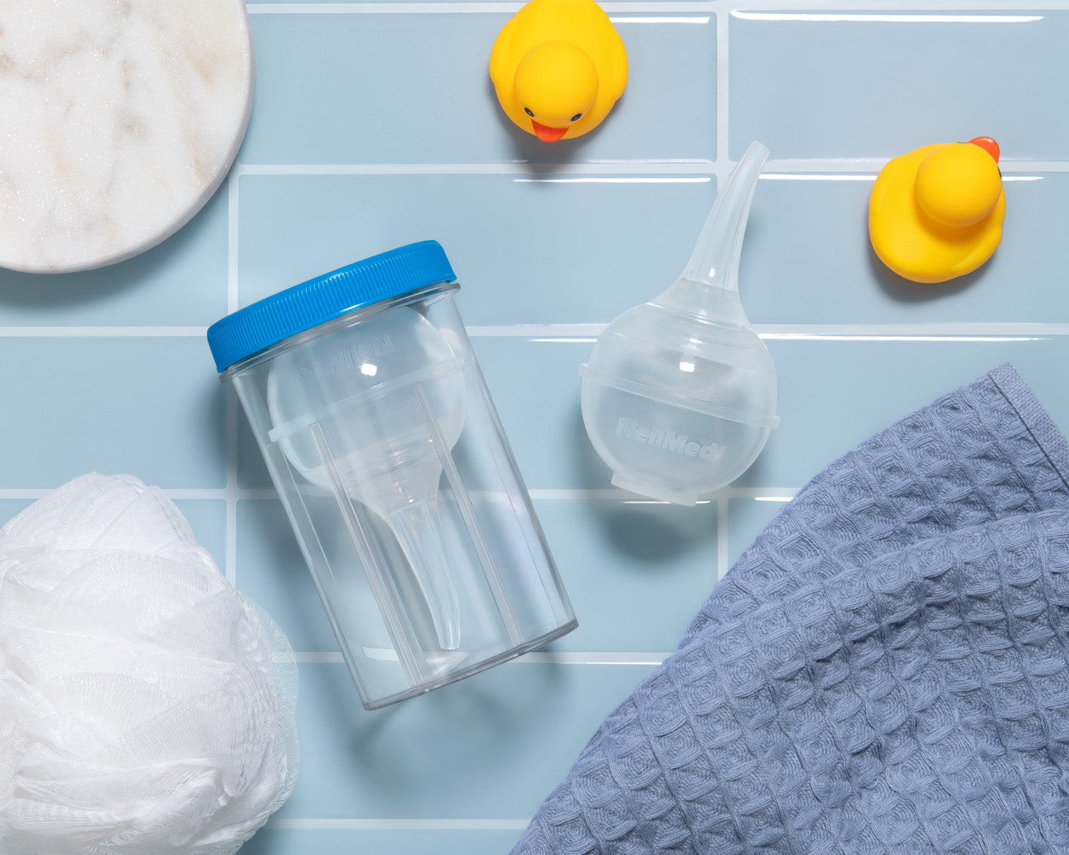 Clear plastic cup with blue lid, funnel, white towel, and rubber duckies on a light blue tiled background